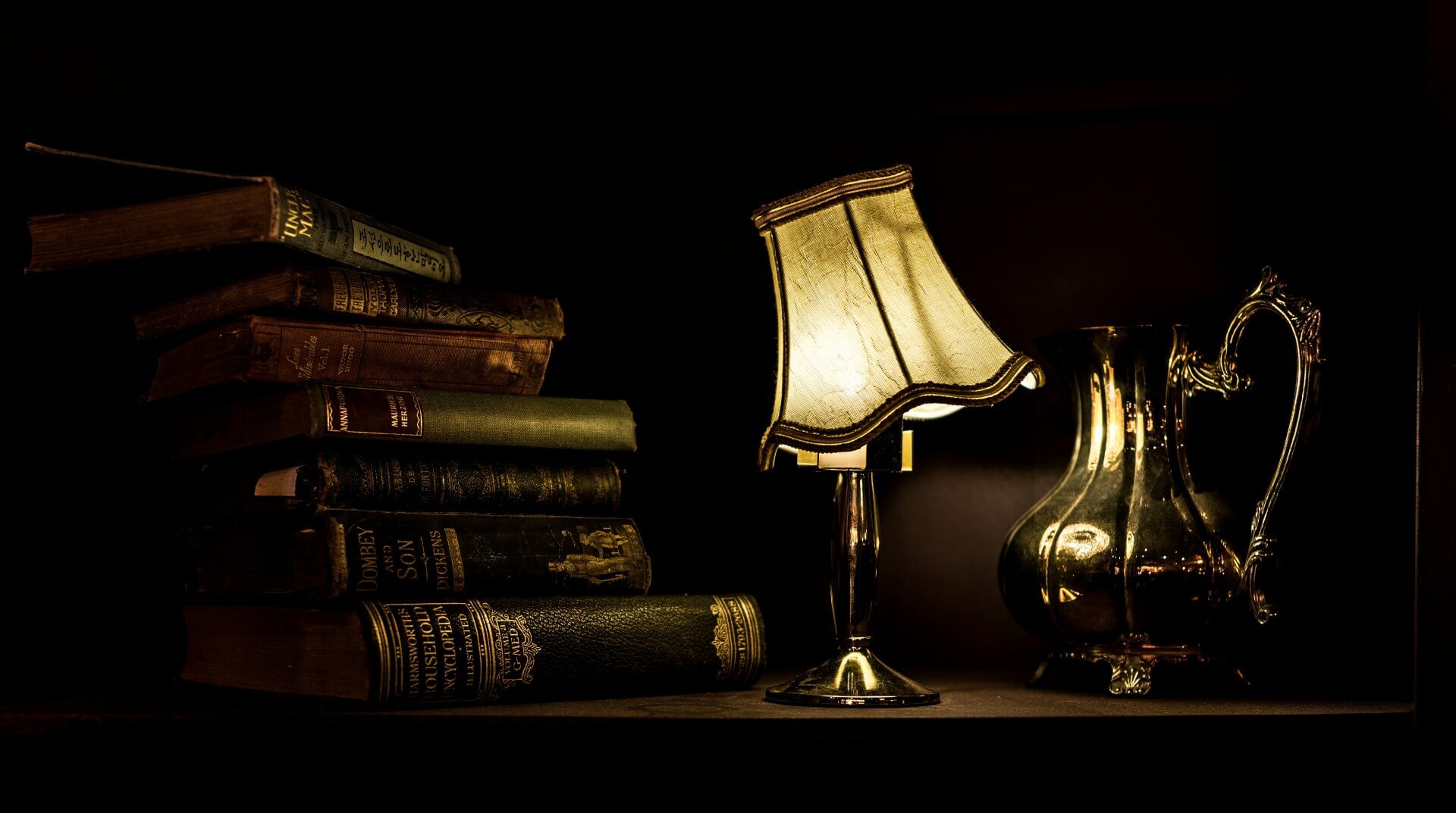dark picture of a stack of books on the left of a desk and a crooked lampshade on a desk lamp on the righthand side